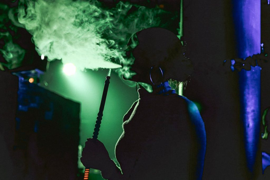 A woman enjoys smoking shisha in a dark, neon-lit environment, exhaling a cloud of smoke.